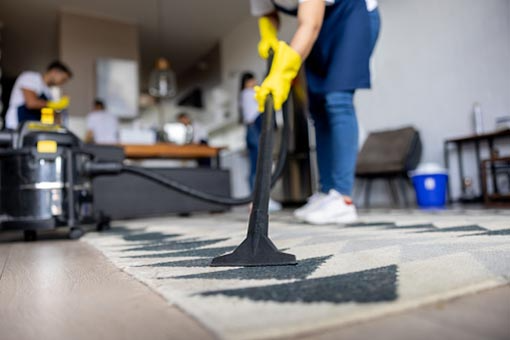 A person wearing yellow gloves is cleaning a patterned rug with a vacuum cleaner in a room with other people.
